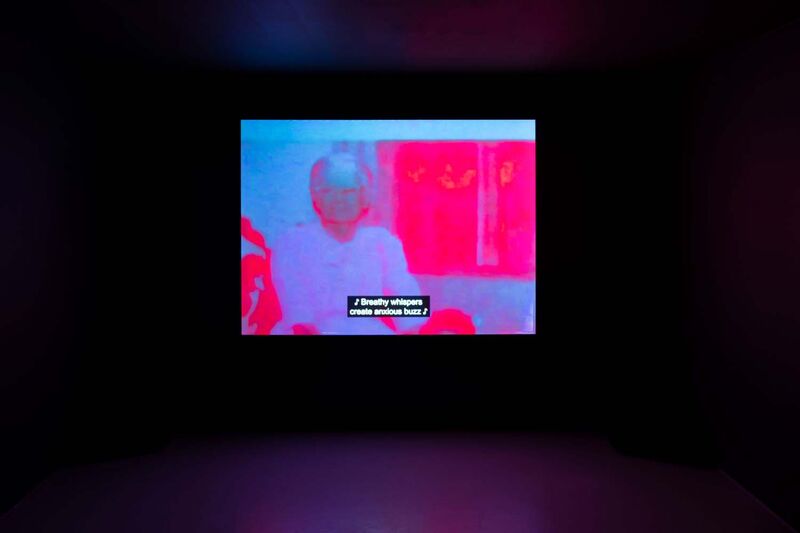 a screen in a dark gallery space with a red. blue and purple toned image of an elderly lady 