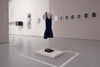 A small child's school uniform a dress hangs from the ceiling in a gallery with other works on show in the background 