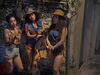 Three feminine people stand leaning against a wall, smoking. One wears a vest and demin shorts, showing tattooed skin. Two have curly hair and cowboy hats on, with fringed demin vests and leather shorts, straddling hobby horses, as though taking a break from performing in a show.