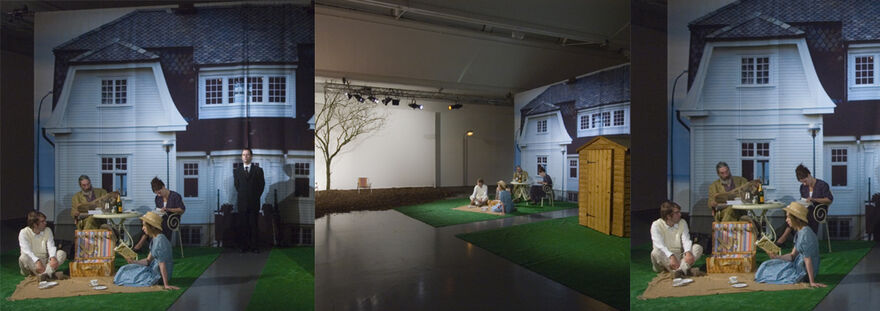 From Killing Time at DCA Galleries. Four people have a picnic on fake gras in the galleries in front of a large fabric sheet with a photograph of a house on it. There is a real wooden shed next to the people.