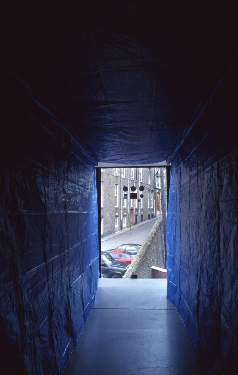 A dark room is covered in blue plastic wrapping, a large open door looks out on to a street and car park.