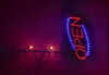 Black barbed metal work is installed on a red painted wall illuminated by pink light. to the left of the image a neon shop light saying 'open' in blue and red is hung on its end. 