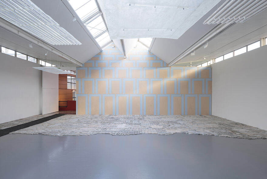 Photograph shows an installation in gallery 2, with natural light. Stright ahead on the back wall is patterned wall coverings with orange blocks in a grid on a greaty background. Sheets of metal painted white hang from the ceiling overhead. On the floor ahead of us at the back are cast bricks. To the left is a black long strip of rubber leading to the auxiliary space.