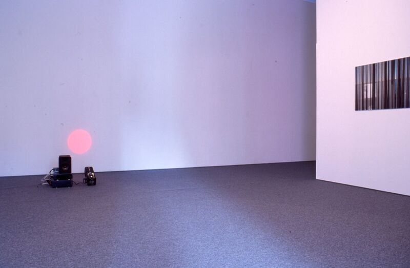 A small projector projects a pink circle onto a white gallery wall