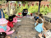 Children sitting around a table in a garden while a DCA artist demonstrates a technique