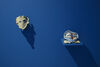 Two sculptural objects akin to masks or costume articles are displayed against a vibrant blue wall. To the left, a beige object has holes cut into it like eyes, it is directly mounted on the wall. To the right, a blue hood lays on a shelf protruding from the wall. 