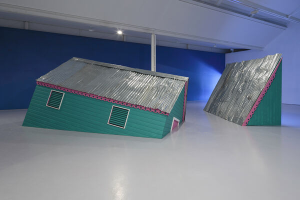 An installation image of gallery two showing a green structure like a building with a corrugated roof and a pink trim, as though it is sinking into the floor.