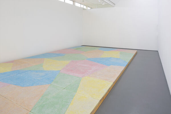 A floor based work is shown in gallery one. The walls are white and the gallery floor is painted grey. On top of this, a sculptural floor is lain in blocks, raising the floor over most of the gallery. This work is brightly coloured, with pastel blue green, yellow orange and pink, in abstract shapes.
