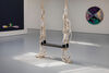 An installation photograph of the artist's work in Gallery 2 at DCA, showing an artwork in the form of a swing, with a dark wood seat made from found materials, and white braided rope. In the background can be seen a round colourful canvas on a white wall. Streams of sunlight hit the grey floor below the swing. 