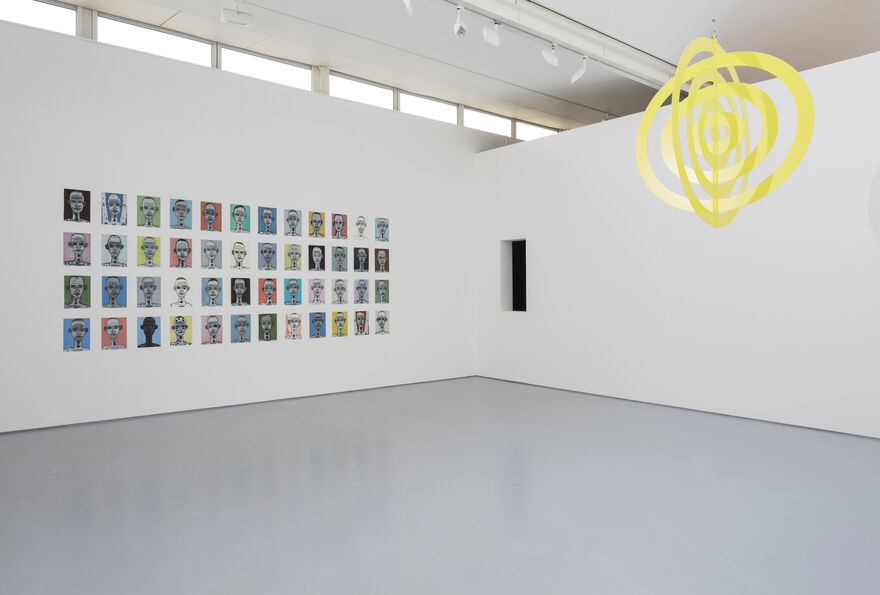 Image shows a hanging yellow artwork to the right, which is strung from the ceiling and consists of flat concentric rings. To the left we can see many small works of art ranging in bright colours which are hung in a grid on the white wall. To the corner of the gallery, in the centre of the image, is a small dark window allowing a view to the adjacent gallery.