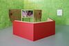 A gallery with green walls. A video screen in the corner showing a figure in red in a cave. A red seating area in front of the screen.