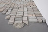 Photograph taken close to the floor, looking along the surface of several cast bricks laid out on the floor in a grid. The brick closest to the camera is crushed.