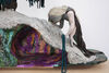 A close up of a textile work on a wooden plinth, with a stooping figure to the right and a cave like structure to the left, the outside of which is grey wool and the inside is painted with purple, orange and shiny green.