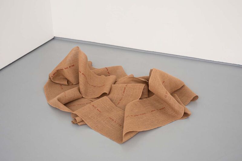 Crumpled heap of jute fabric in the corner of a gallery with red words visible