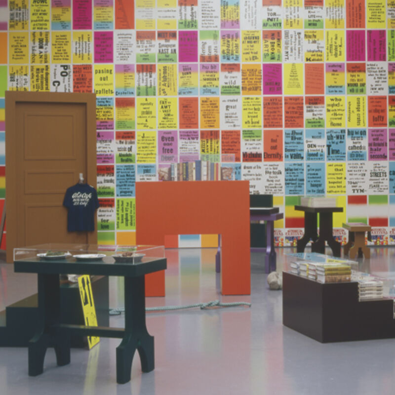 From Allen Ruppersberg's exhibition at DCA. The wall is covered in colourful, graphic posters with slogans on them. There are also small black tables which are covered with plates and books. There is a brown door leaning against the poster wall, and this has a t-shirt hanging on the door handle.